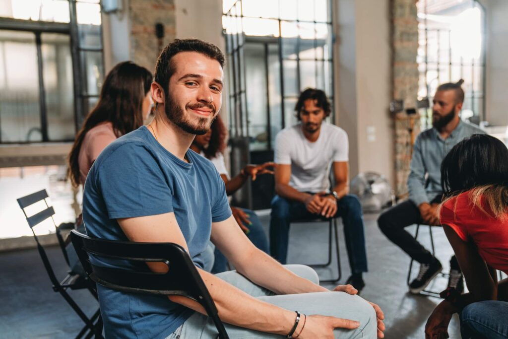 Portrait of a man looking at camera during a group therapy session. Mental health concept in a loft with big windows. Healthcare and medicine concept. Group of people sitting in a circle are participating in a support meeting.